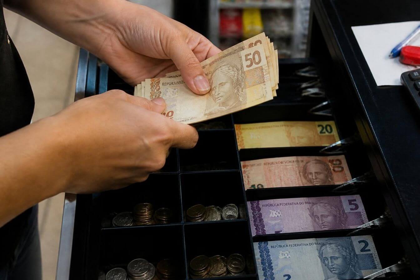 Mãos contando notas de real na gaveta do caixa
